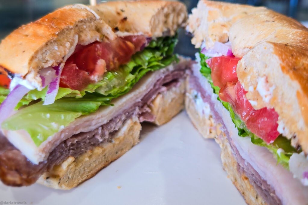 Close-up of a sliced bagel sandwich filled with roast beef, turkey, lettuce, tomato, and onion
