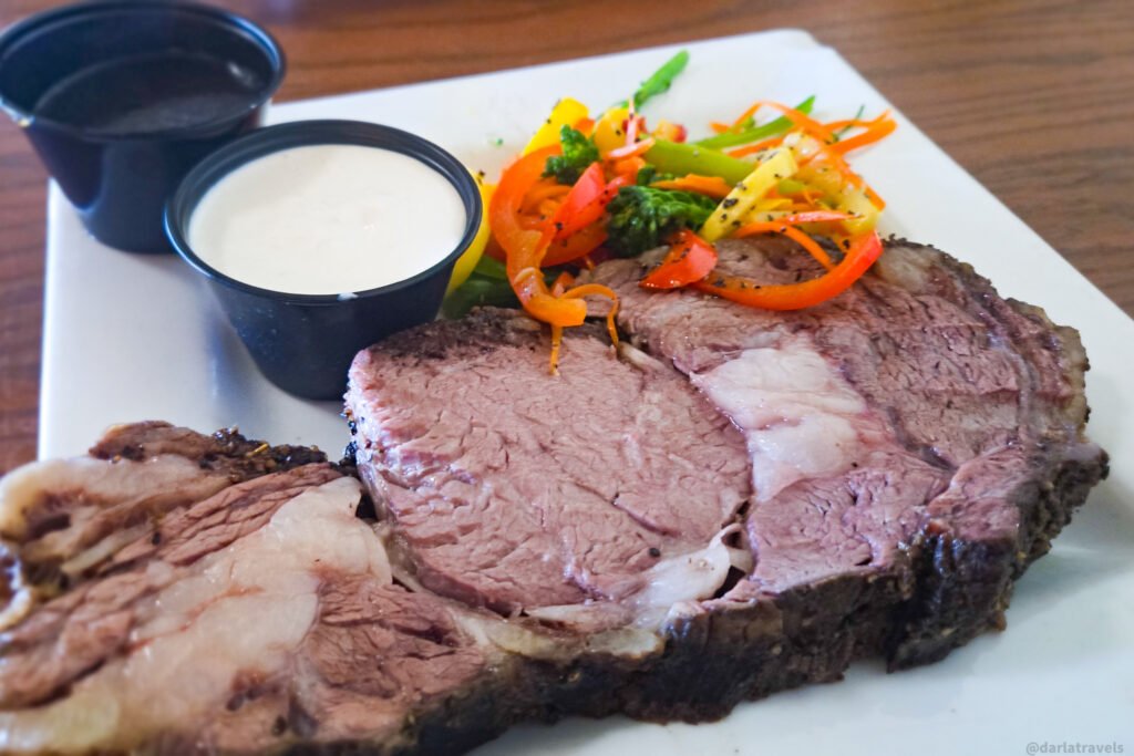 A white plate holds sliced prime rib, colorful vegetables, and two cups of sauce.