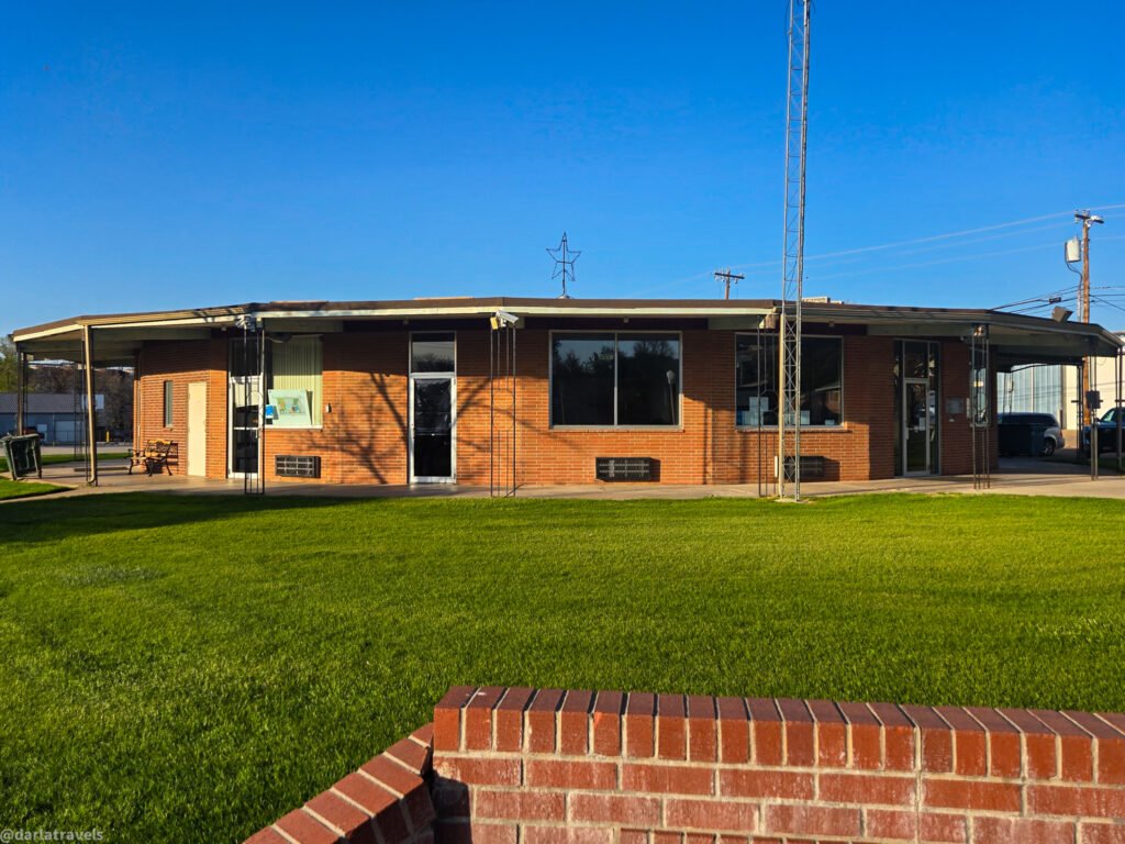 A low, brick building with a covered walkway and a tall radio tower under a clear blue sky.