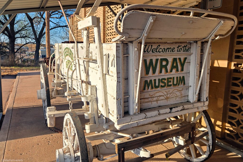 An antique white wagon with the words "Welcome to the WRAY MUSEUM" painted on the back is parked outside on a concrete walkway.