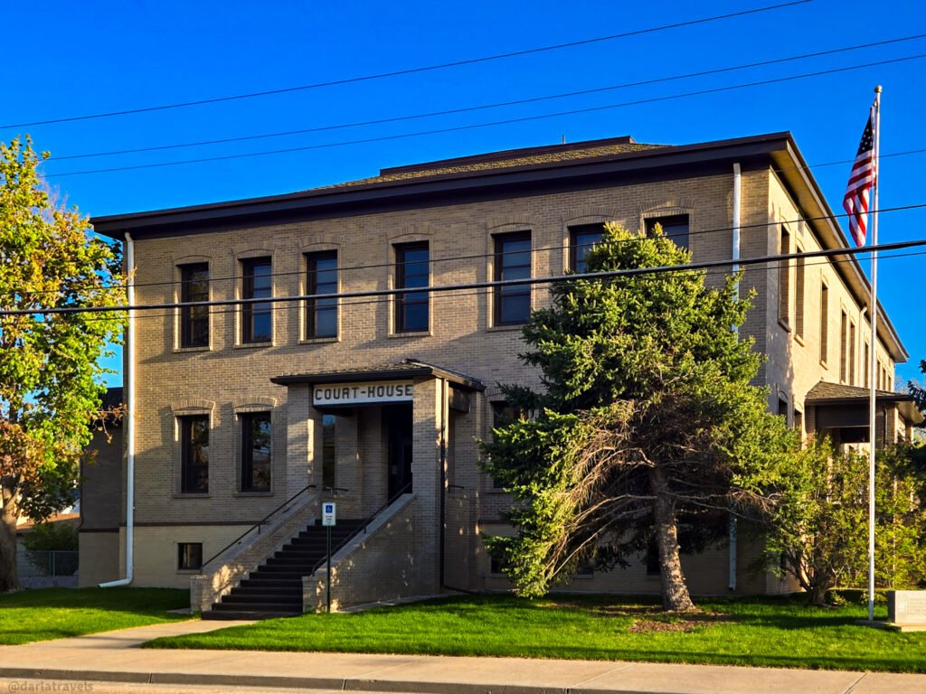 A light brick courthouse building with a US flag flying on the right side under a clear blue sky.