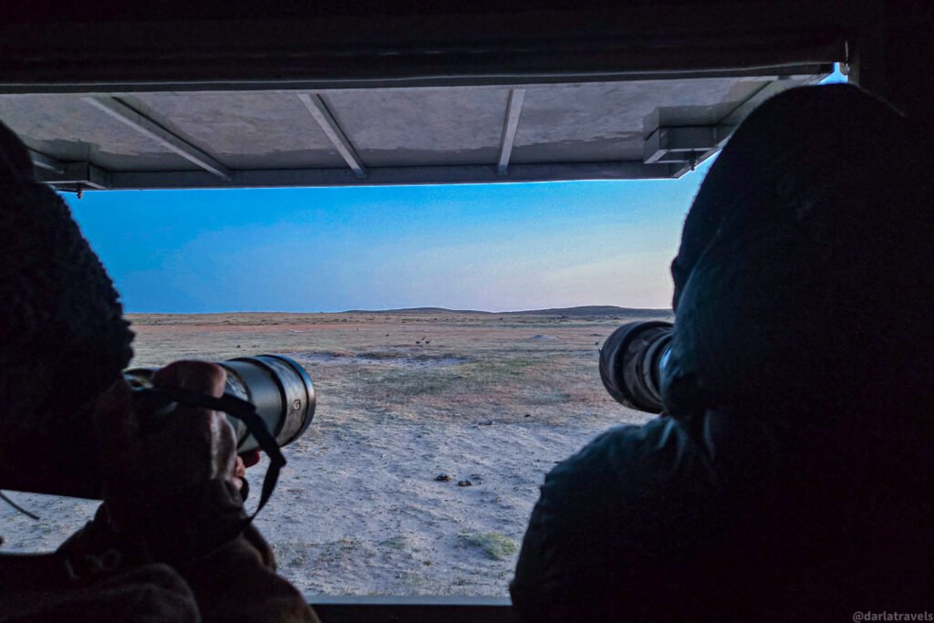 Silhouetted photographers look out from a bird viewing trailer, aiming telephoto lenses at an expansive, arid landscape under a clear blue sky.