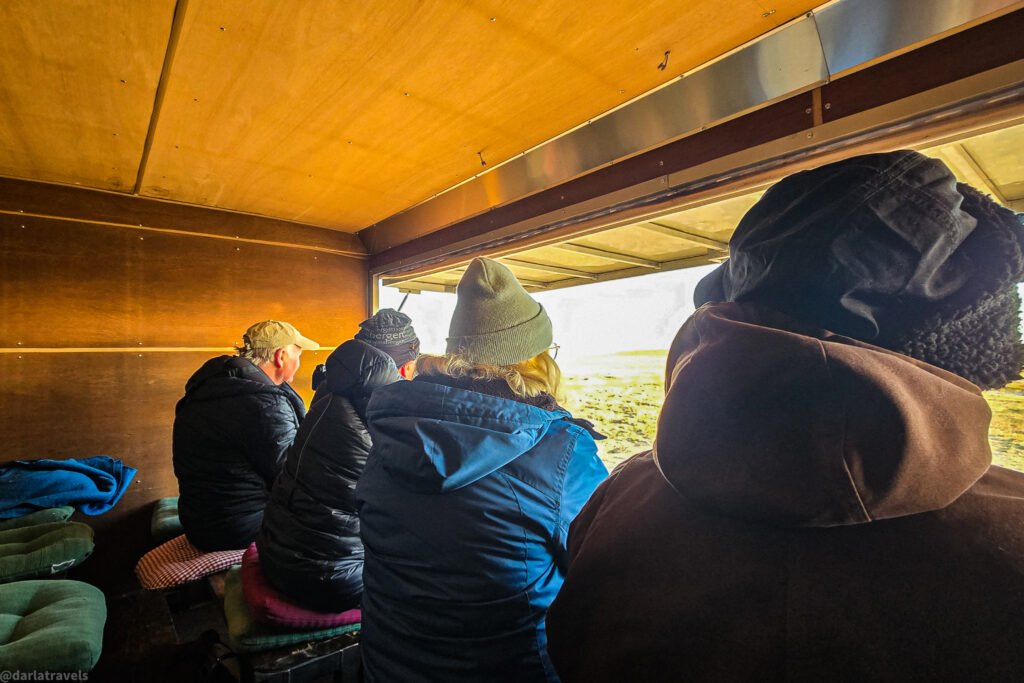 People wearing winter clothing sit inside a bird viewing tailer with an open side, looking out at a landscape.