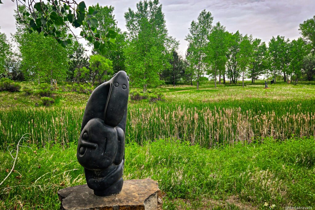 Stylized dark stone sculpture on a low pedestal surrounded by tall grass and leafy trees.