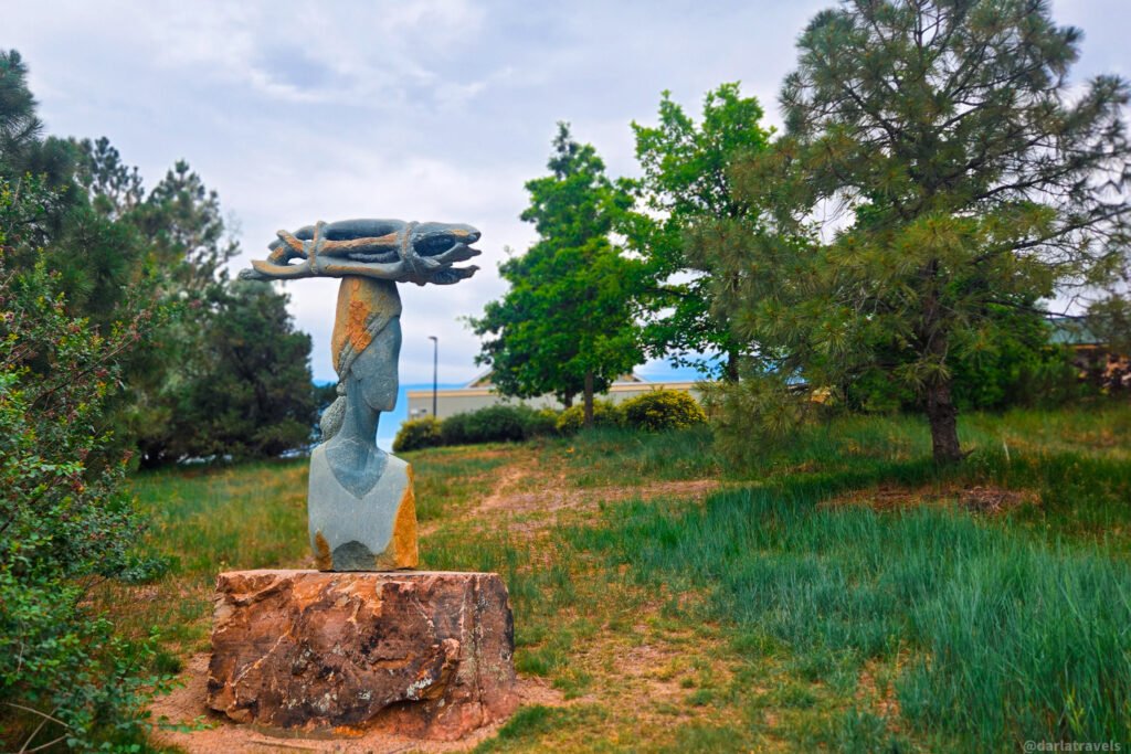 Elongated stone figure with a rounded head and top bundle set on a rough stone base in a grassy area.