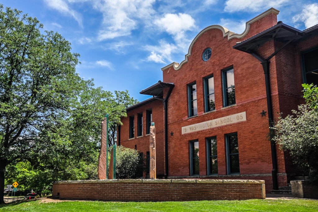 Red-brick Washington School building with “Washington School ’05” carved above the windows and three tall abstract sculptures on the lawn in Loveland, Colorado.
