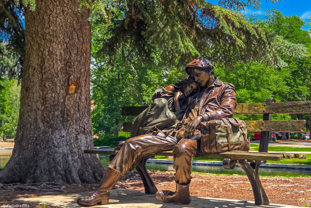 Two bronze figures seated close together on a wooden bench under a large tree near a pond.