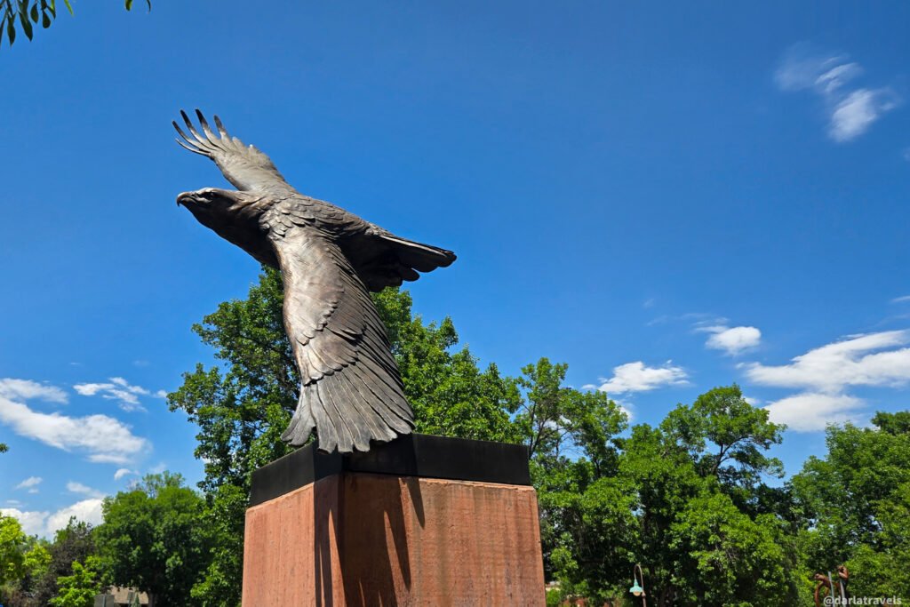 Bronze eagle with outstretched wings mounted on a tall stone pedestal against a blue sky.