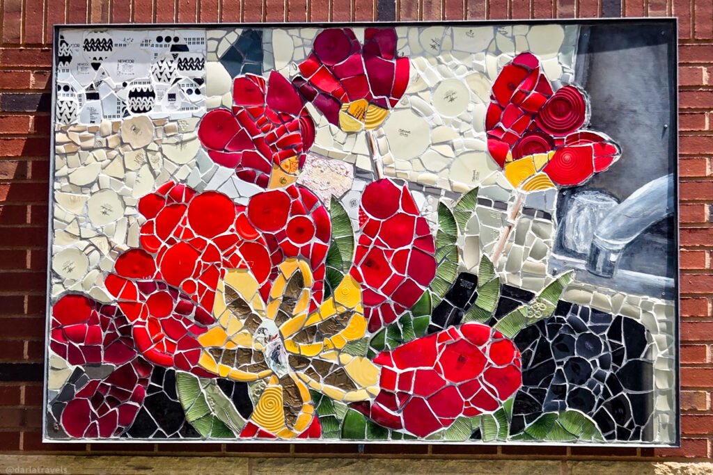 Mosaic wall panel of red, yellow, and green flowers made from broken ceramic pieces, mounted on a red-brick wall in Loveland, Colorado.