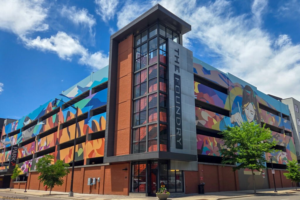 Brick and metal parking garage with colorful murals and a tall glass stair tower under a partly cloudy sky