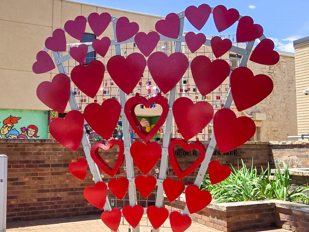 Metal tree sculpture with red heart shapes and a wall of colorful padlocks in a sunny courtyard.
