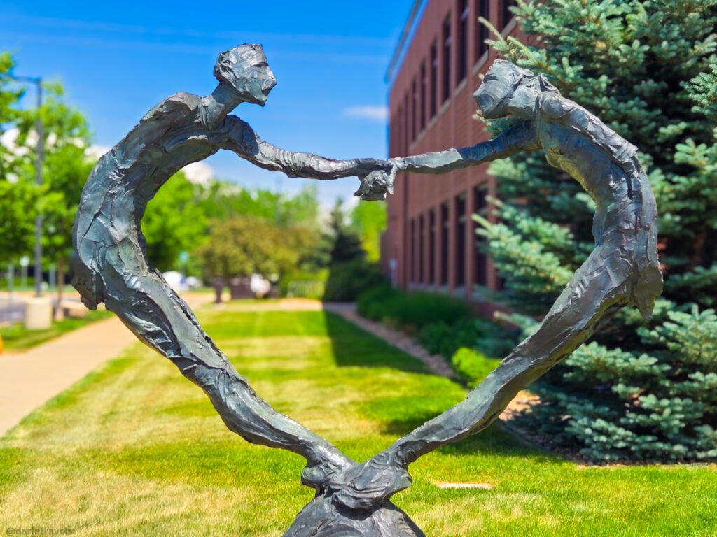 Two elongated bronze figures reaching together to form a heart shape on a grassy outdoor lawn.