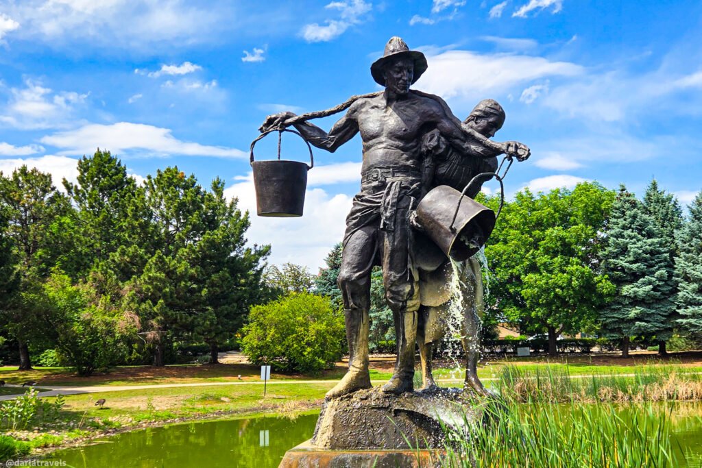 Bronze figures with buckets on a stone base, one pouring water into a pond surrounded by greenery.