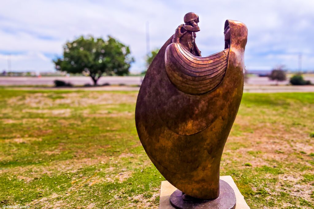 Curved bronze figure enveloping a smaller figure on a concrete pedestal in a grassy outdoor setting.