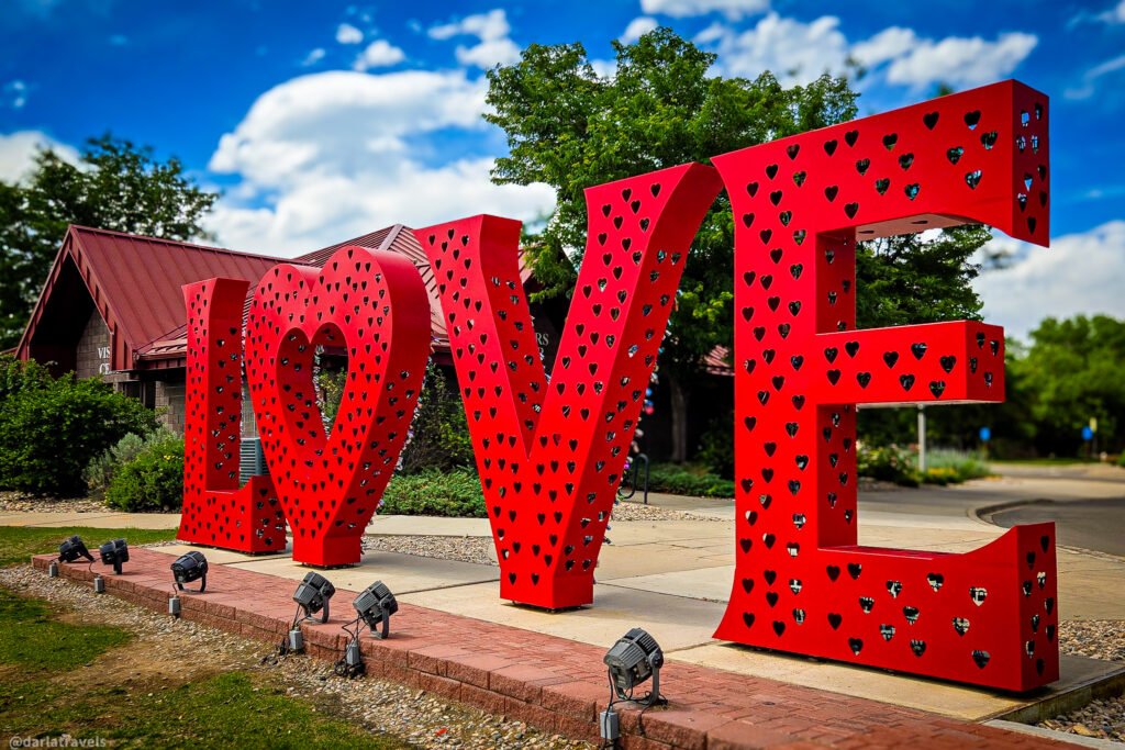 Large red metal “LOVE” sculpture with heart-shaped cutouts, covered with colorful padlocks, on a landscaped plaza in Loveland, Colorado.