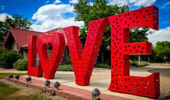 Large red metal “LOVE” sculpture with heart-shaped cutouts, covered with colorful padlocks, on a landscaped plaza in Loveland, Colorado.