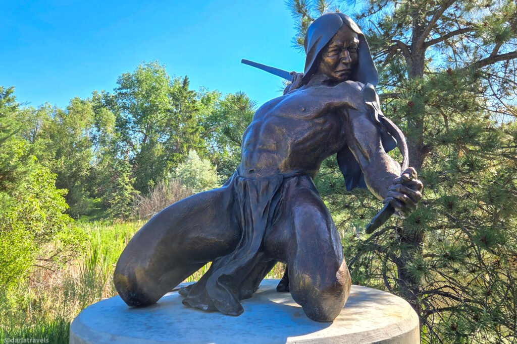 Bronze sculpture of a kneeling, hooded figure testing a bow, set beside reeds and pine trees in Loveland, Colorado.