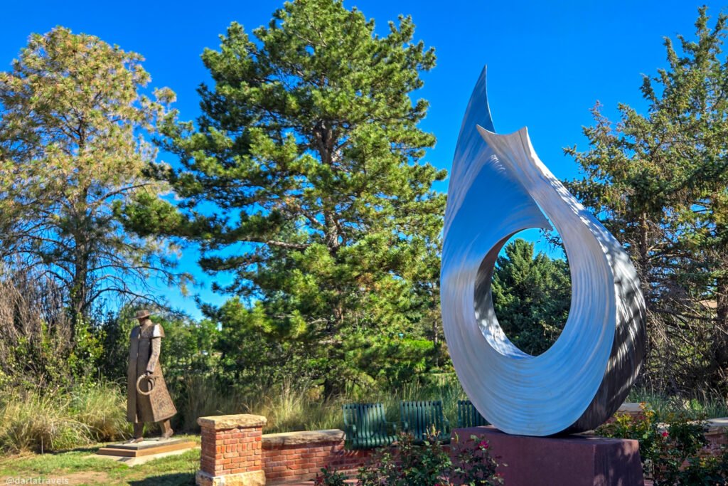Tall silver abstract metal sculpture with a circular opening, set among trees and benches, with a bronze figure in the background in Loveland, Colorado.