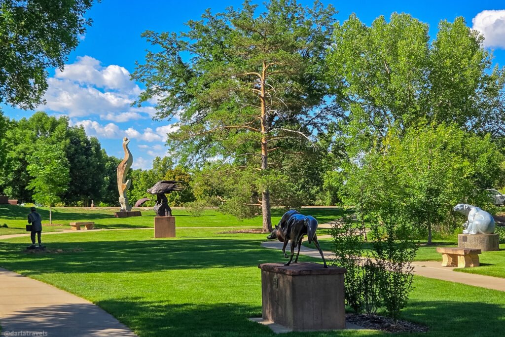 Sculptures among green lawns and mature trees at Benson Sculpture Garden in Loveland, Colorado, beneath a bright blue sky.