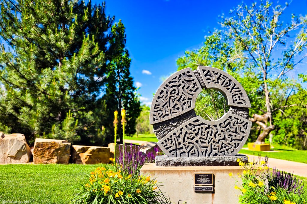 Circular stone sculpture with carved patterns and a center opening on a pedestal in a landscaped garden.