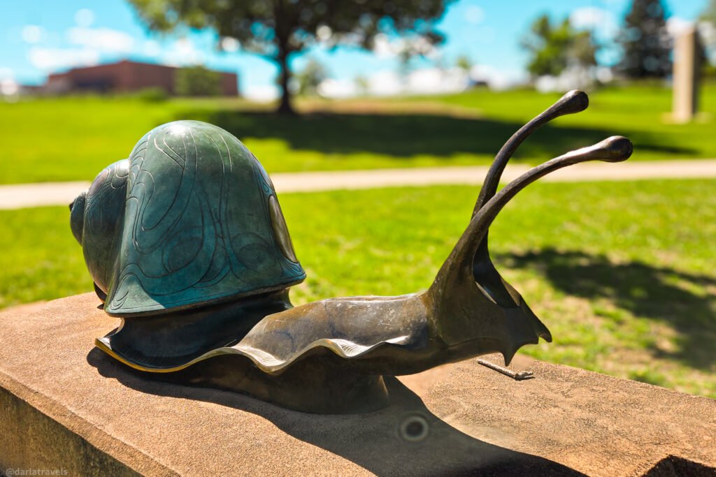 Bronze snail sculpture with a patterned green shell and long curved antennae on a low stone wall beside a grassy path in Loveland, Colorado.