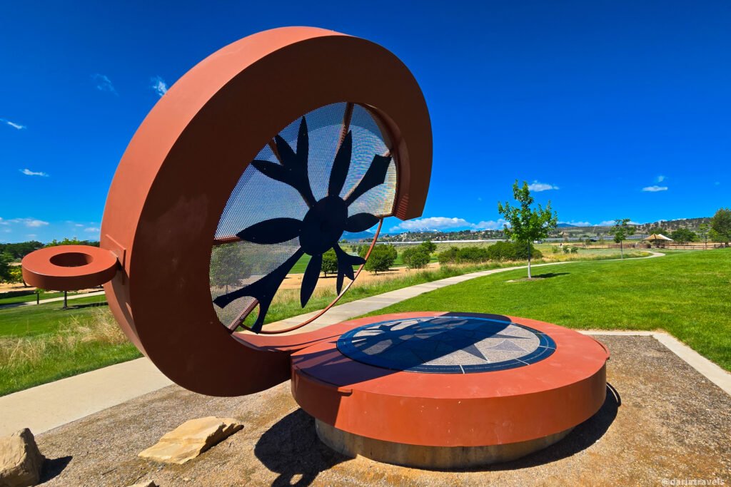 Large rust-colored metal sculpture with a circular frame, black windmill-like cutout, and compass design on a round base beside a paved path in Loveland, Colorado.
