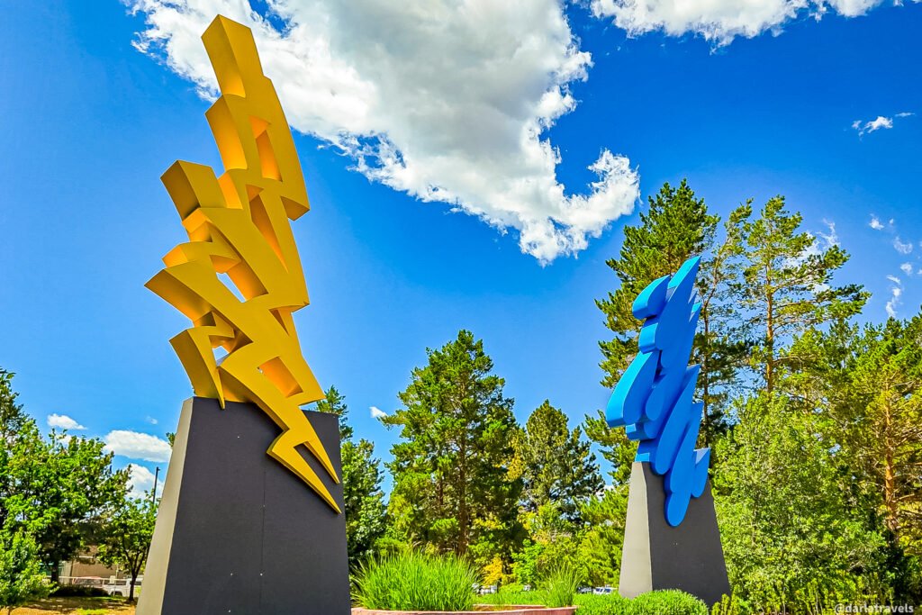 Two tall abstract metal sculptures—one yellow and one blue—rise from dark pedestal bases in a landscaped Loveland park.