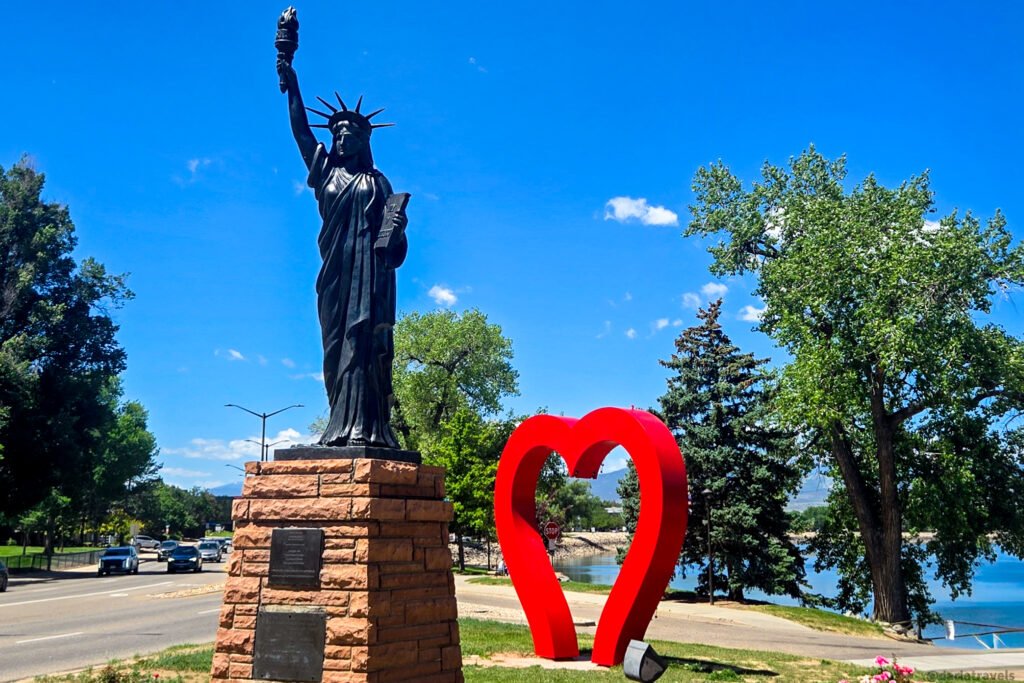 Statue of Liberty replica beside a bright red heart sculpture near a waterfront park, with trees, mountains, and blue sky in Loveland, Colorado.
