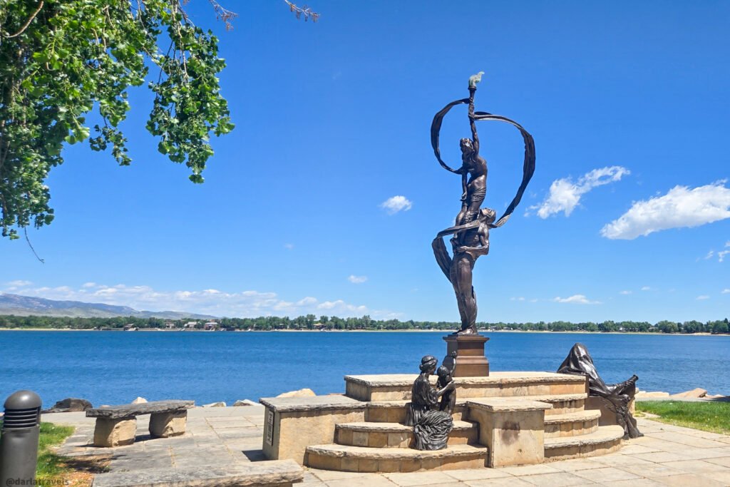 Bronze sculpture of two figures reaching upward around a looping ribbon form, with smaller seated figures on stone steps beside a lake in Loveland, Colorado.