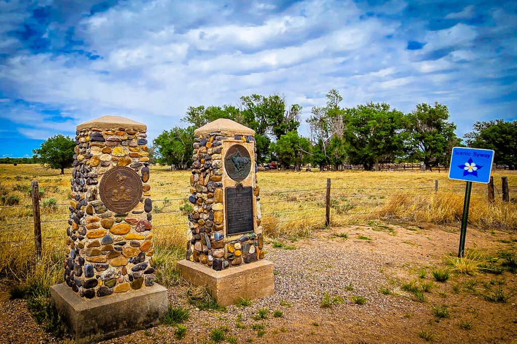 Old Julesburg stone monuments with bronze plaques beside a fenced field and a Scenic Byway sign.