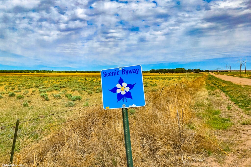 Blue Scenic Byway sign with a columbine emblem beside a gravel road through open plains.