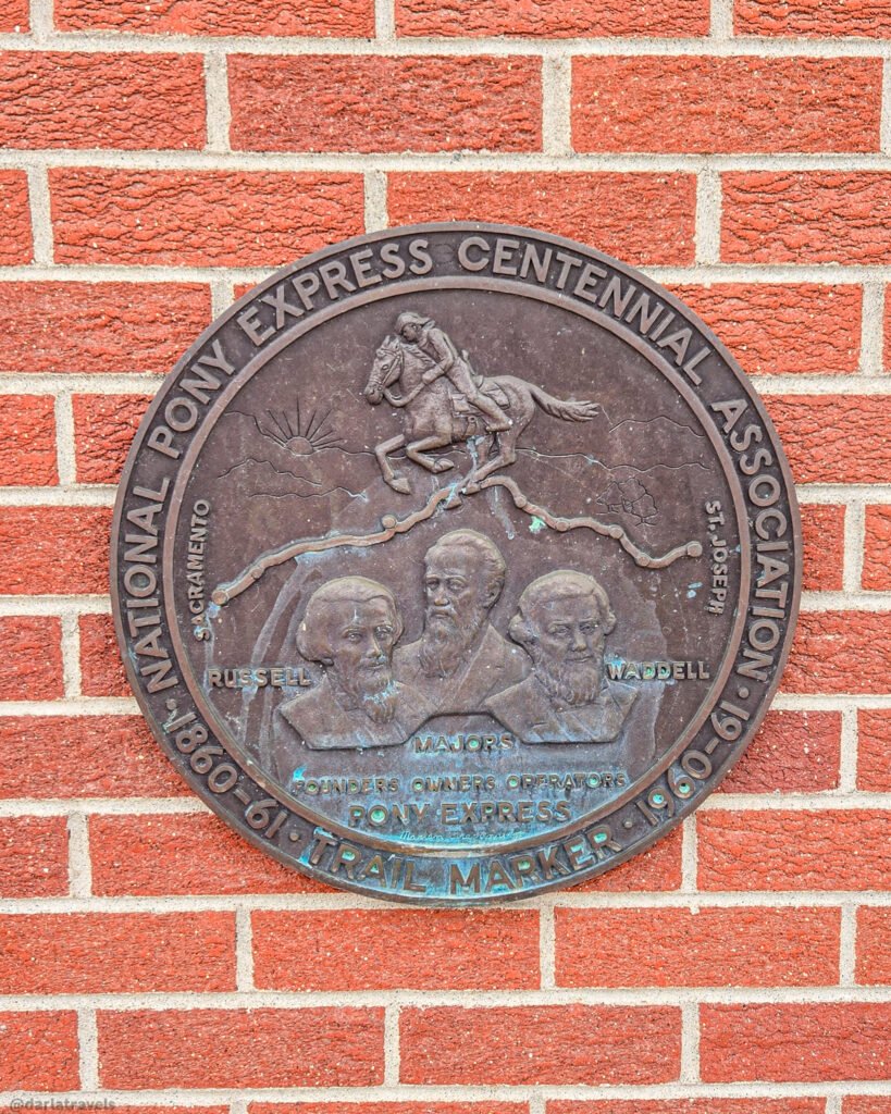 Circular Pony Express centennial trail-marker plaque on a red-brick wall, with a mounted rider, route illustration, and portraits labeled Russell, Majors, and Waddell.