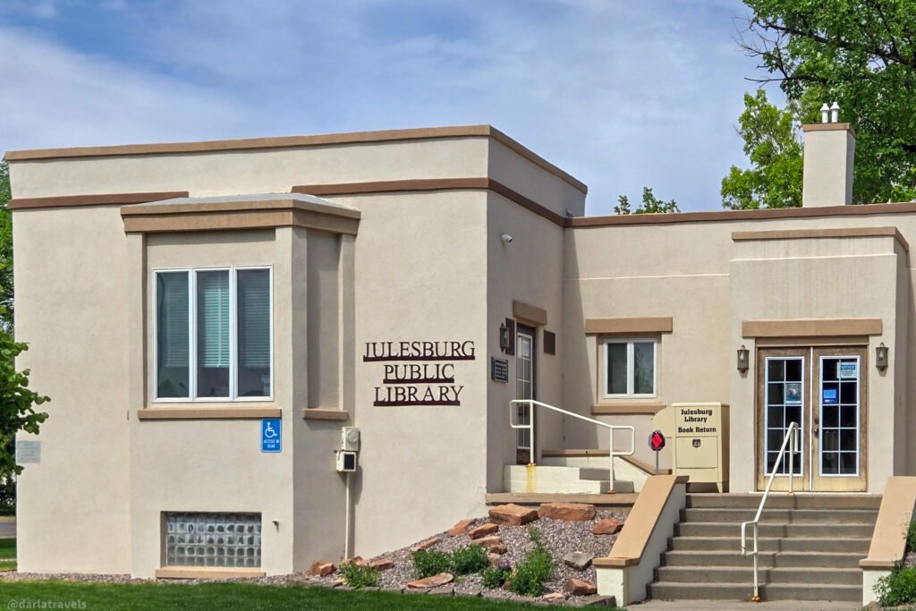 Julesburg Public Library exterior with tan stucco walls, entrance steps, and a book return box.