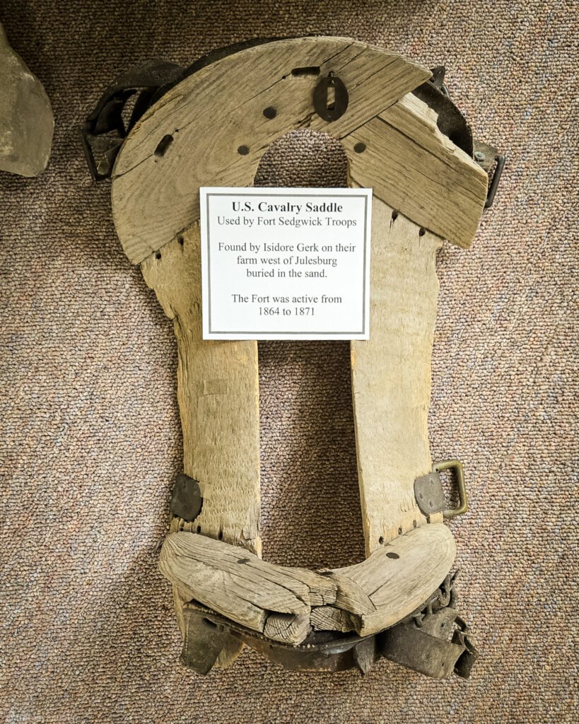 Weathered wooden saddle framework with metal fittings and a label identifying it as a U.S. cavalry saddle used by Fort Sedgwick troops.