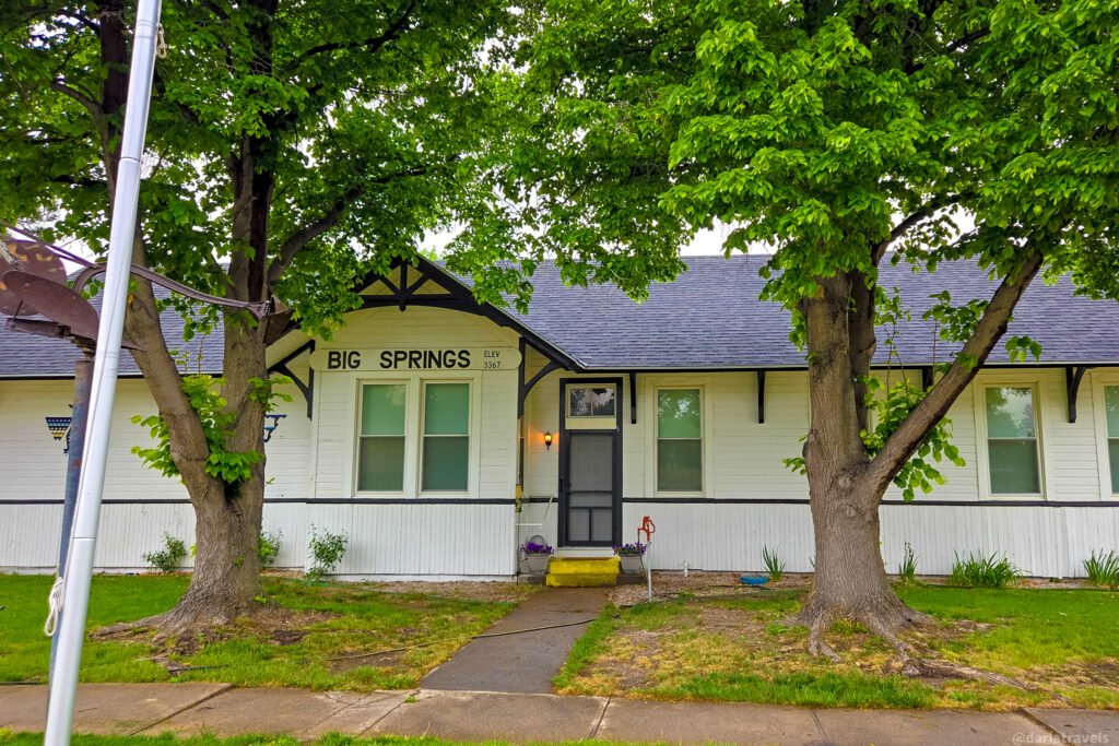 Big Springs Depot Museum’s white exterior beneath leafy trees, with a station-name sign and yellow entrance steps.
