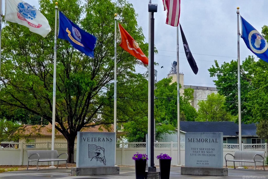 Big Springs veterans memorial with military flags, two inscribed stone panels, benches, and flower planters.