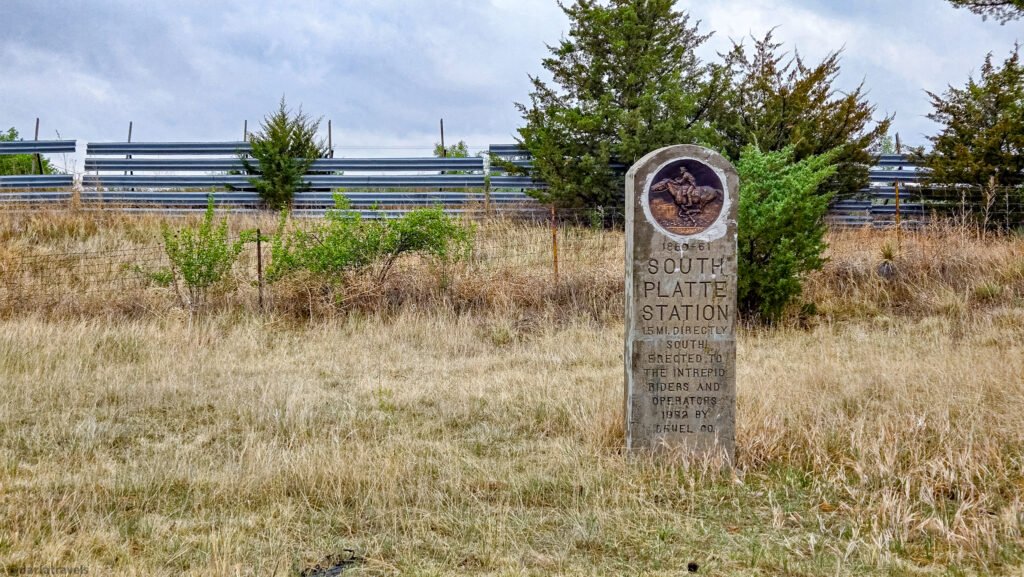 South Platte Station Pony Express marker with a rider medallion, standing in dry grass beside a wire fence.
