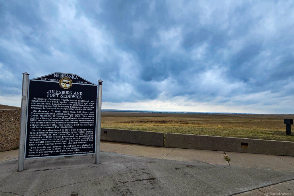 Nebraska historical marker titled “Julesburg and Fort Sedgwick,” overlooking open plains beneath dark clouds.