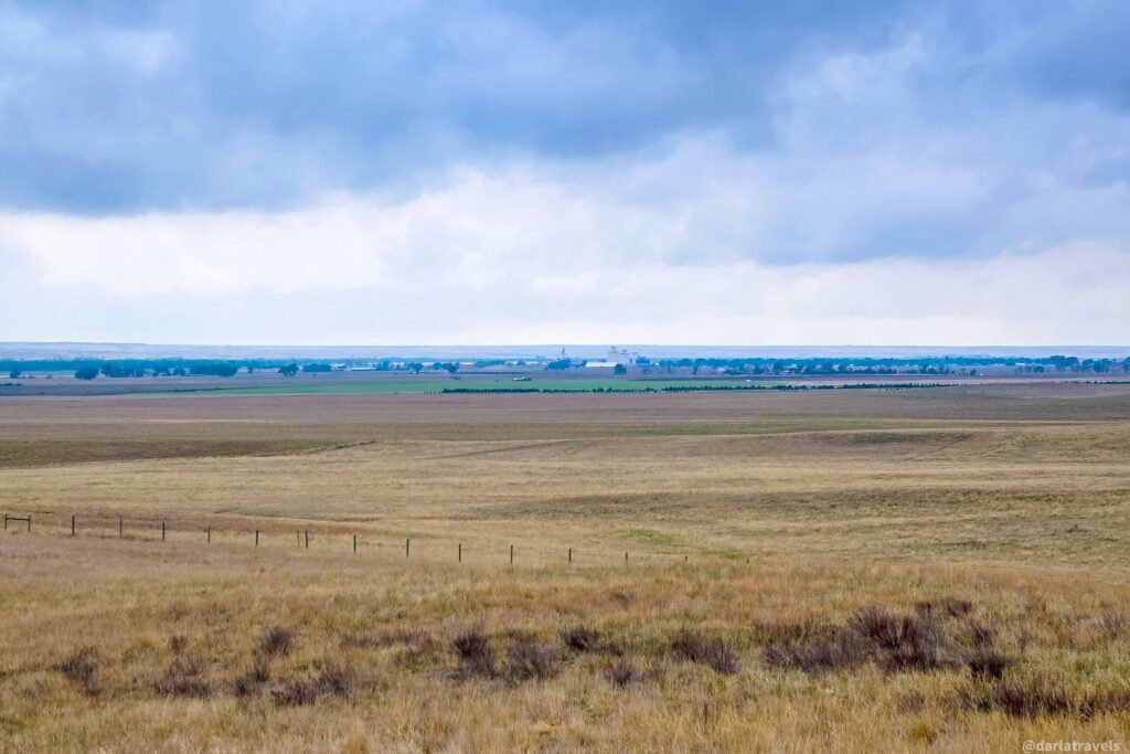 Open grassland and farm fields stretching toward distant buildings beneath a cloudy sky.