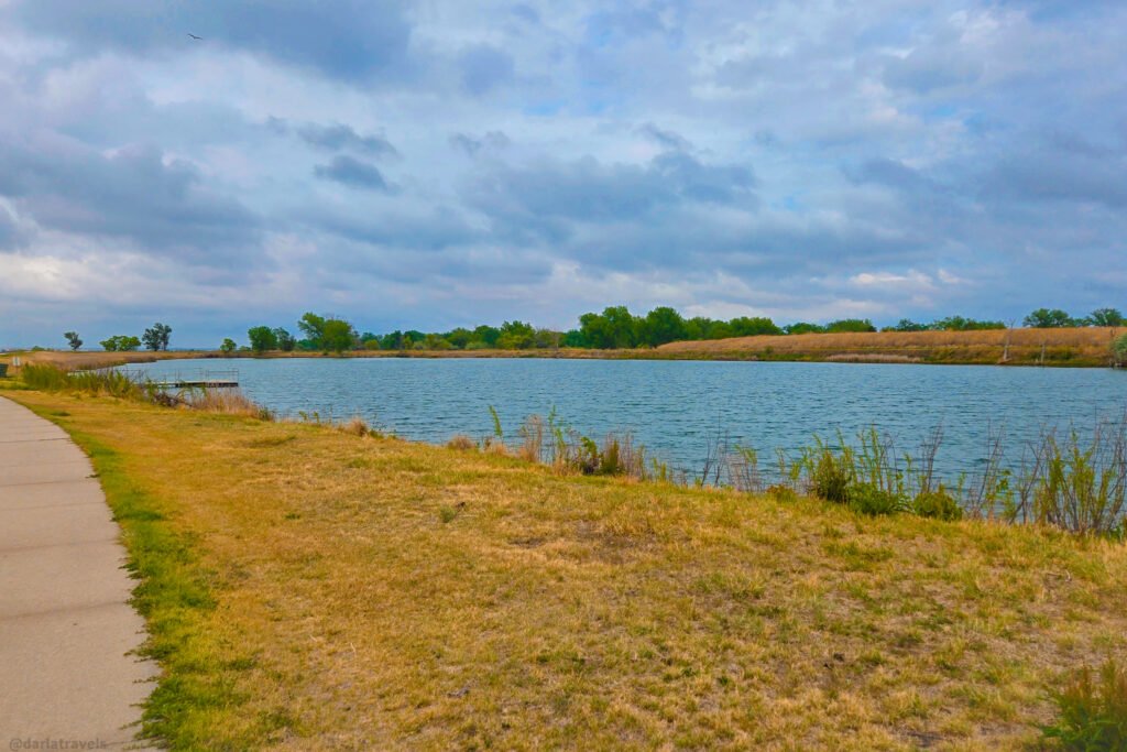 DePoorter Lake in Julesburg, with a paved shoreline path, a small dock, and reeds along the water.